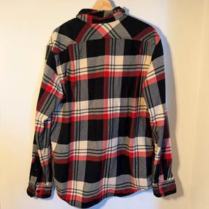 以下が含まれることがあります： 黒、赤、白、グレーのクラシックなパターンが特徴のチェック柄フランネルシャツ。長袖のシャツはボタンダウンで、袖口はボタン留め。木製のハンガーにかかっています。
