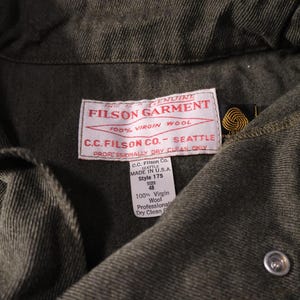 Puede incluir: Primer plano de una prenda Filson verde oscuro. La etiqueta dice "FILSON GARMENT 100% VIRGIN WOOL C.C. FILSON CO. - SEATTLE". La prenda tiene cierre de bot&oacute;n y una etiqueta que indica que est&aacute; hecha en EE. UU., estilo 175, talla 48.