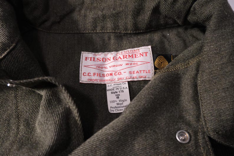 Puede incluir: Primer plano de una prenda Filson verde oscuro. La etiqueta dice "FILSON GARMENT 100% VIRGIN WOOL C.C. FILSON CO. - SEATTLE". La prenda tiene cierre de bot&oacute;n y una etiqueta que indica que est&aacute; hecha en EE. UU., estilo 175, talla 48.