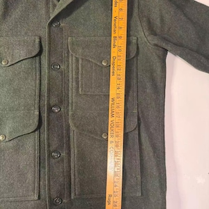 May include: A dark green wool jacket with four front pockets and button closures. The jacket has a collared neckline and a tag on the upper left. A wooden ruler is placed next to the jacket, measuring approximately 29 inches.