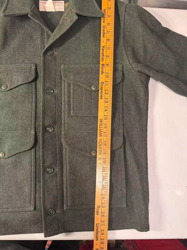 May include: A dark green wool jacket with four front pockets and button closures. The jacket has a collared neckline and a tag on the upper left. A wooden ruler is placed next to the jacket, measuring approximately 29 inches.