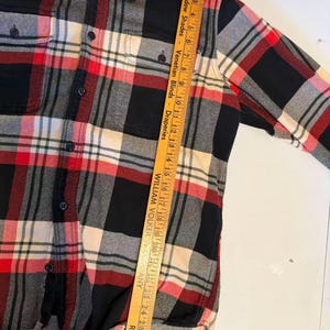以下が含まれることがあります： 黒、赤、クリーム色のチェック柄フランネルシャツ。胸ポケットが2つ付いています。シャツはボタンダウンフロントと襟付きです。「FILSON」のブランド名がラベルに表示されています。木製の定規でシャツを測定しています。