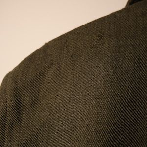 Puede incluir: Primer plano de una tela verde oliva oscura, posiblemente un abrigo o una chaqueta. El material tiene un aspecto texturizado con un tejido diagonal visible y algunas imperfecciones menores. Se ve la zona del hombro, lo que sugiere una prenda.