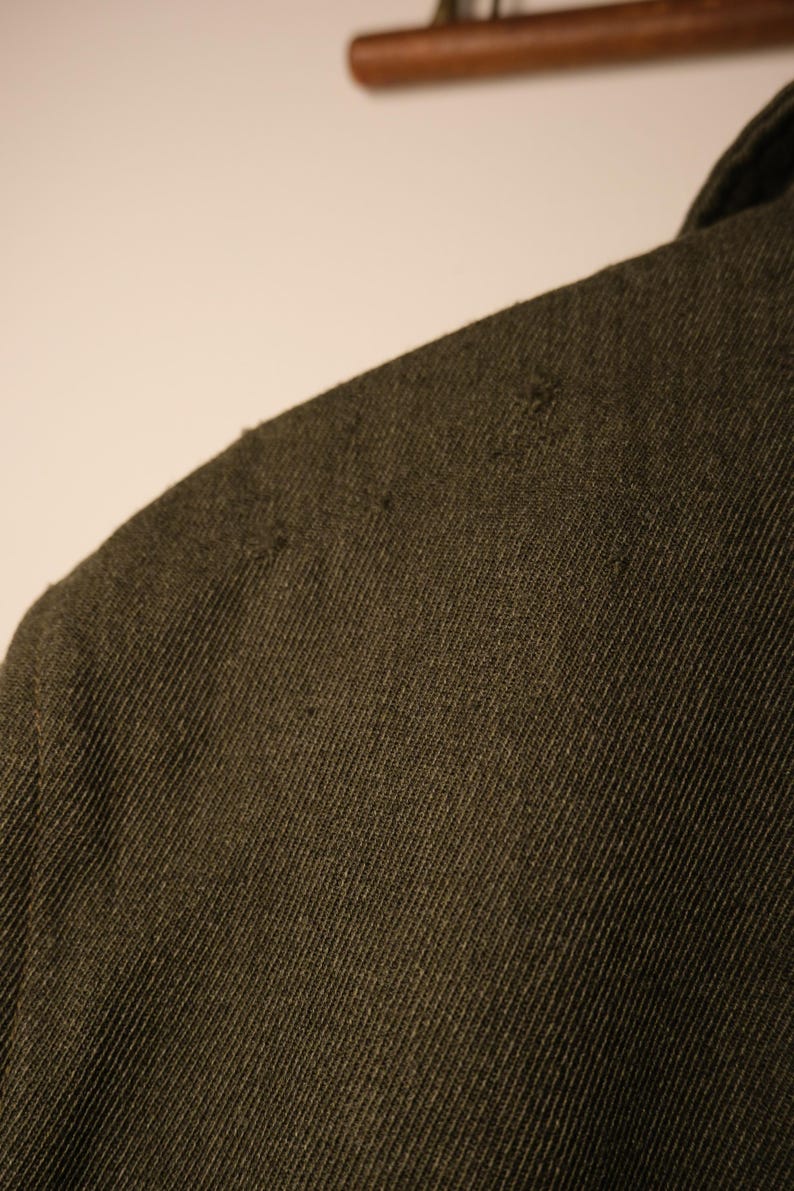 Puede incluir: Primer plano de una tela verde oliva oscura, posiblemente un abrigo o una chaqueta. El material tiene un aspecto texturizado con un tejido diagonal visible y algunas imperfecciones menores. Se ve la zona del hombro, lo que sugiere una prenda.