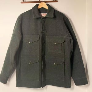 May include: A dark gray wool jacket with four front pockets and button closures. The jacket has a collared neckline and long sleeves. The garment is hanging on a wooden hanger against a white wall.