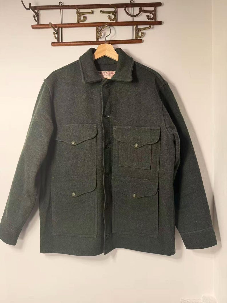 May include: A dark gray wool jacket with four front pockets and button closures. The jacket has a collared neckline and long sleeves. The garment is hanging on a wooden hanger against a white wall.