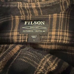 May include: Close-up of a dark brown and tan plaid shirt. The shirt has a Filson label that reads "FILSON SINCE 1897 C.C. FILSON CO. - SEATTLE, WA MADE IN INDIA L". The shirt is made of a soft, woven fabric.