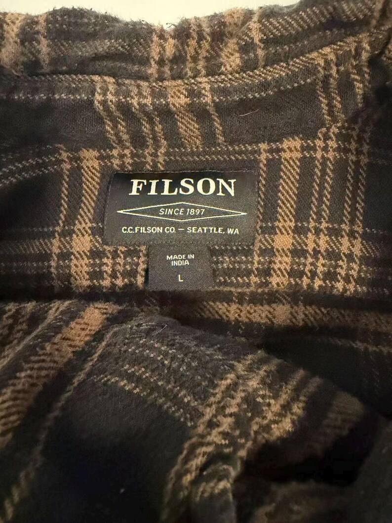May include: Close-up of a dark brown and tan plaid shirt. The shirt has a Filson label that reads "FILSON SINCE 1897 C.C. FILSON CO. - SEATTLE, WA MADE IN INDIA L". The shirt is made of a soft, woven fabric.