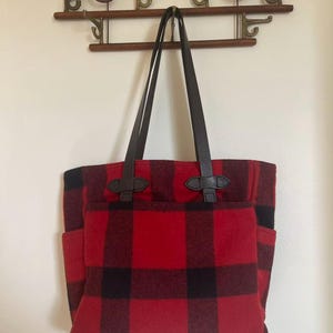 Op de afbeelding: Rood-zwarte buffelruit tote bag met bruin lederen handvatten en accenten. De tas heeft twee buitenvakken en hangt aan een houten wandrek. De tas is gemaakt van een dik, duurzaam materiaal.