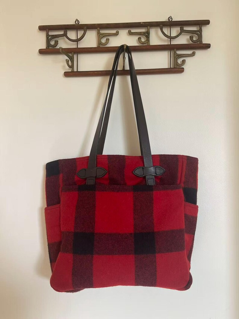 Op de afbeelding: Rood-zwarte buffelruit tote bag met bruin lederen handvatten en accenten. De tas heeft twee buitenvakken en hangt aan een houten wandrek. De tas is gemaakt van een dik, duurzaam materiaal.