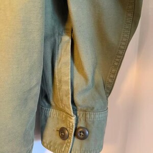 May include: Close-up of an olive green jacket sleeve. The cuff features two dark brown buttons. The fabric has visible stitching and a structured appearance. The jacket appears to be made of a durable material.