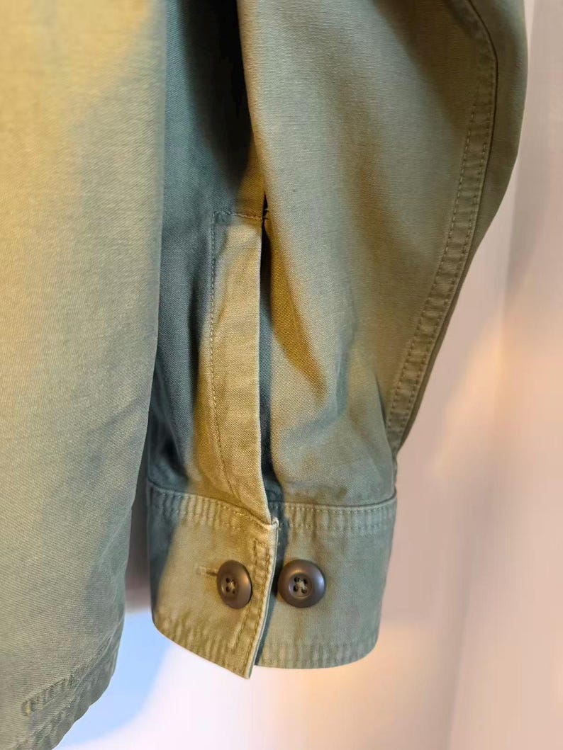 May include: Close-up of an olive green jacket sleeve. The cuff features two dark brown buttons. The fabric has visible stitching and a structured appearance. The jacket appears to be made of a durable material.