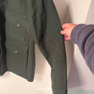 May include: Dark green wool jacket with two chest pockets and button closures. The jacket has a classic design with a tailored fit. The sleeves are long with buttoned cuffs. The jacket is made of a thick, warm material.