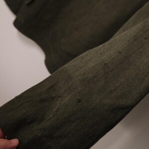 Puede incluir: Primer plano de una manga de chaqueta verde oliva oscuro. La tela tiene un aspecto texturizado con desgaste visible y peque&ntilde;as imperfecciones. La manga est&aacute; en &aacute;ngulo, mostrando el tejido y las variaciones de color de la tela.