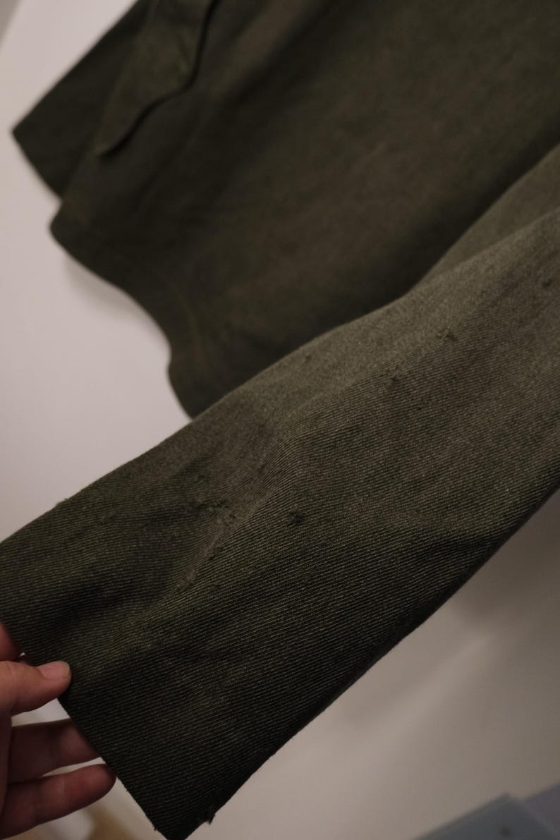 Puede incluir: Primer plano de una manga de chaqueta verde oliva oscuro. La tela tiene un aspecto texturizado con desgaste visible y peque&ntilde;as imperfecciones. La manga est&aacute; en &aacute;ngulo, mostrando el tejido y las variaciones de color de la tela.