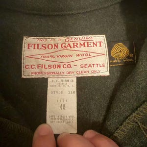 May include: Close-up of a dark green Filson garment. The label reads "FILSON GARMENT 100% VIRGIN WOOL C.C. FILSON CO. - SEATTLE". The size tag indicates size 40. The garment is made of 100% virgin wool.