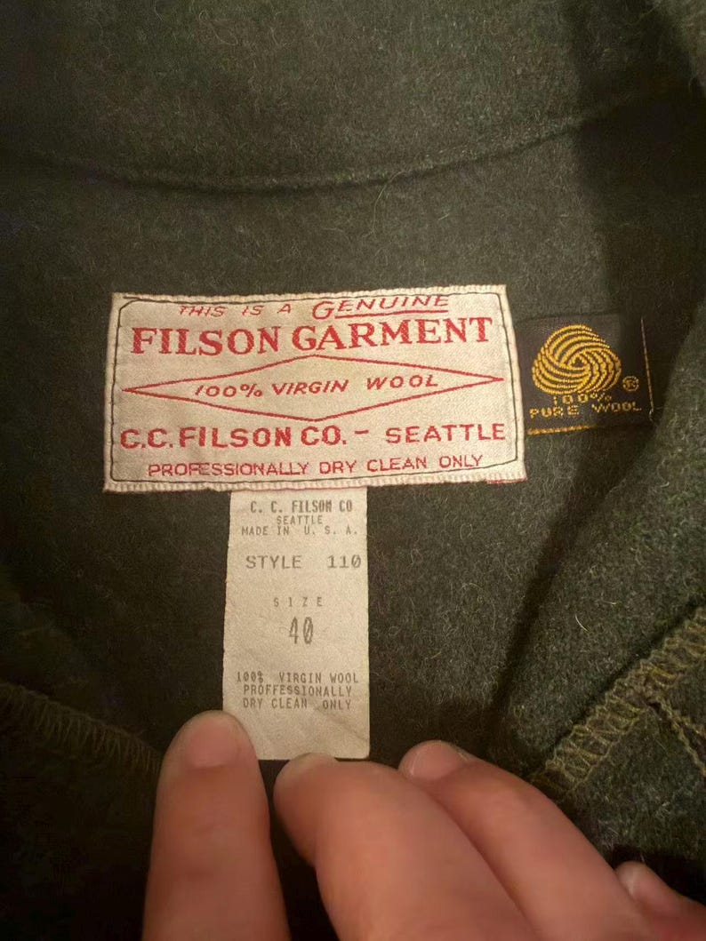 May include: Close-up of a dark green Filson garment. The label reads "FILSON GARMENT 100% VIRGIN WOOL C.C. FILSON CO. - SEATTLE". The size tag indicates size 40. The garment is made of 100% virgin wool.