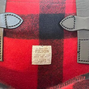 Op de afbeelding: Close-up van een rood en zwart geruite stof met leren riemen. De stof heeft een gedurfd, klassiek patroon. Een klein label leest "A Genuine Filson" met de tekst "Might as well have the best."
