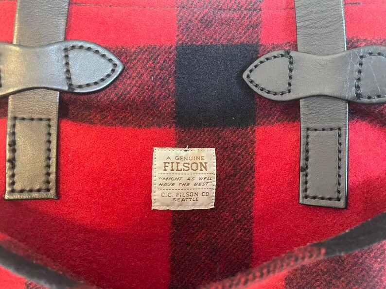 Op de afbeelding: Close-up van een rood en zwart geruite stof met leren riemen. De stof heeft een gedurfd, klassiek patroon. Een klein label leest "A Genuine Filson" met de tekst "Might as well have the best."
