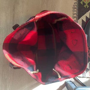 Op de afbeelding: Een rood-zwarte geruite draagtas met bruine leren handvatten. De binnenkant van de tas is zichtbaar en toont een grote capaciteit. De tas is gemaakt van een dik, geweven materiaal en is ontworpen voor het dragen van spullen.