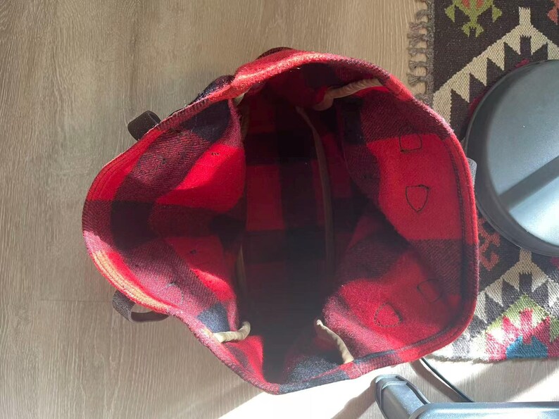 Op de afbeelding: Een rood-zwarte geruite draagtas met bruine leren handvatten. De binnenkant van de tas is zichtbaar en toont een grote capaciteit. De tas is gemaakt van een dik, geweven materiaal en is ontworpen voor het dragen van spullen.