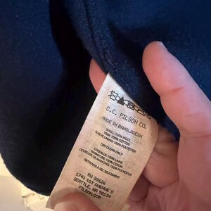 Puede incluir: Prenda azul marino con etiqueta "C.C. FILSON CO." y detalles de materiales: 100% lana para el exterior, 100% algod&oacute;n para el ribete y 100% poli&eacute;ster para el forro de la manga. Fabricado en Bangladesh.