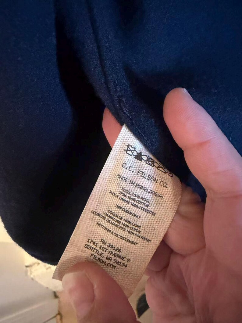 Puede incluir: Prenda azul marino con etiqueta "C.C. FILSON CO." y detalles de materiales: 100% lana para el exterior, 100% algod&oacute;n para el ribete y 100% poli&eacute;ster para el forro de la manga. Fabricado en Bangladesh.