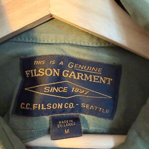 May include: Close-up of a green Filson garment shirt with a navy blue label. The label reads "THIS IS A GENUINE FILSON GARMENT SINCE 1897 C.C. FILSON CO. - SEATTLE". The shirt is size M and made in Sri Lanka.