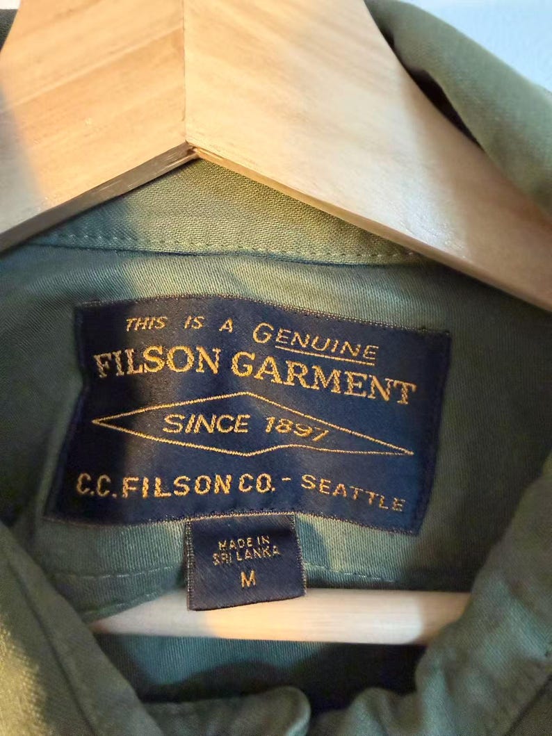 May include: Close-up of a green Filson garment shirt with a navy blue label. The label reads "THIS IS A GENUINE FILSON GARMENT SINCE 1897 C.C. FILSON CO. - SEATTLE". The shirt is size M and made in Sri Lanka.