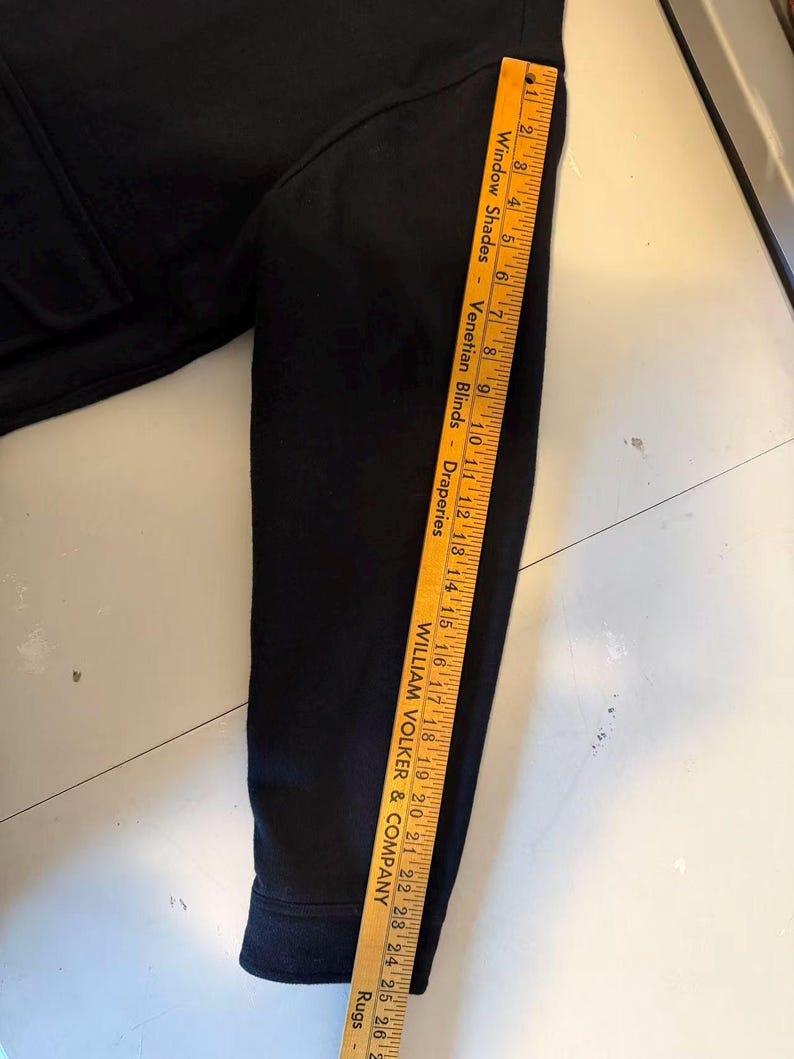 Puede incluir: Una prenda negra de manga larga con una regla colocada a lo largo de la manga. La regla es amarilla con marcas y texto en negro. La prenda parece estar hecha de un material suave.