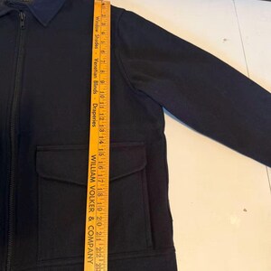 Puede incluir: Chaqueta azul marino con cremallera completa, dos bolsillos delanteros y cuello. La chaqueta se mide con una regla de madera, mostrando una longitud de aproximadamente 63,5 cm.