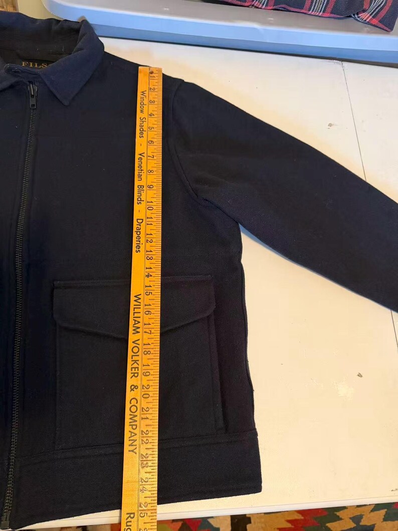 Puede incluir: Chaqueta azul marino con cremallera completa, dos bolsillos delanteros y cuello. La chaqueta se mide con una regla de madera, mostrando una longitud de aproximadamente 63,5 cm.