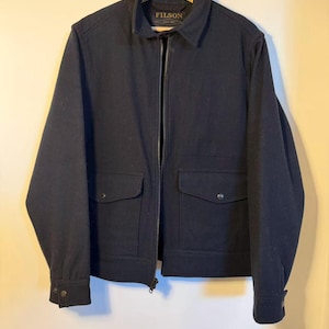 Puede incluir: Chaqueta azul marino con cierre de cremallera completo, dos bolsillos delanteros y pu&ntilde;os abotonados. La chaqueta tiene cuello y la marca "FILSON" es visible.