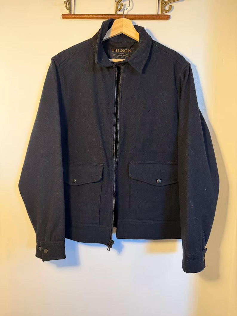 Puede incluir: Chaqueta azul marino con cierre de cremallera completo, dos bolsillos delanteros y pu&ntilde;os abotonados. La chaqueta tiene cuello y la marca "FILSON" es visible.
