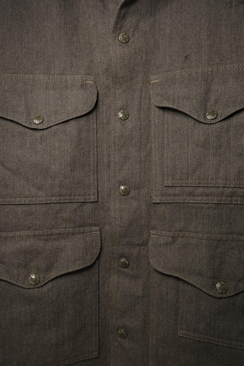 Puede incluir: Primer plano de una camisa abotonada marr&oacute;n con cuatro bolsillos delanteros y cuello puntiagudo. La camisa tiene una tela texturizada y varios botones redondos en el centro. Los bolsillos tienen esquinas redondeadas y est&aacute;n asegurados con peque&ntilde;os botones.