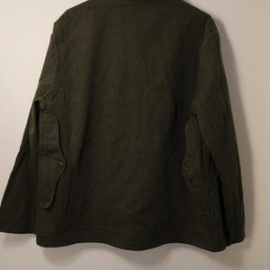 Puede incluir: Chaqueta verde oscuro con cuello y dos bolsillos delanteros. La chaqueta est&aacute; colgada en un perchero de madera contra una pared blanca. La chaqueta tiene cierre de bot&oacute;n y tejido texturizado.