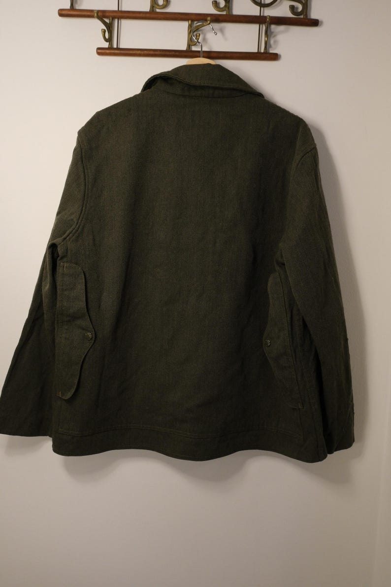 Puede incluir: Chaqueta verde oscuro con cuello y dos bolsillos delanteros. La chaqueta est&aacute; colgada en un perchero de madera contra una pared blanca. La chaqueta tiene cierre de bot&oacute;n y tejido texturizado.