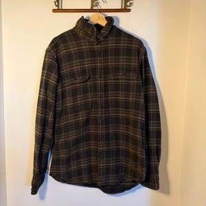 May include: A long-sleeved, button-down shirt in a black and brown plaid pattern. The shirt has two chest pockets and a collared neckline. The garment is hanging on a wooden hanger against a neutral wall.
