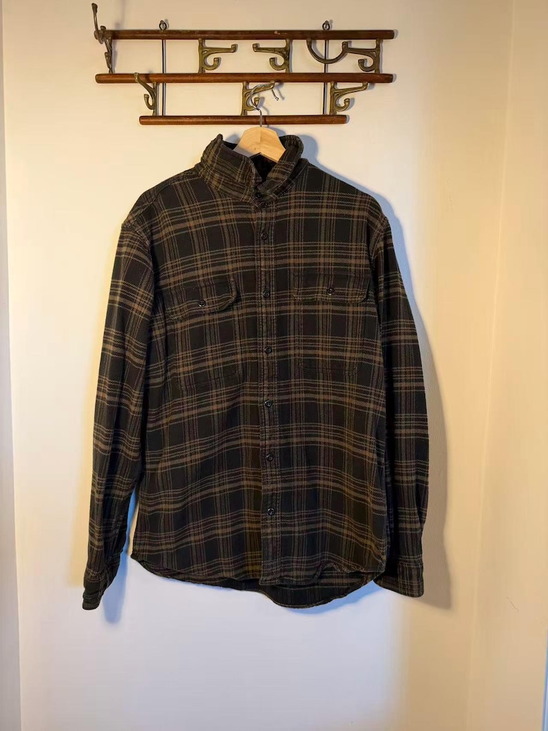 May include: A long-sleeved, button-down shirt in a black and brown plaid pattern. The shirt has two chest pockets and a collared neckline. The garment is hanging on a wooden hanger against a neutral wall.