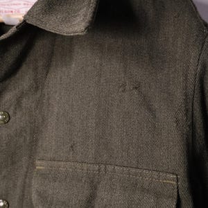Puede incluir: Primer plano de una camisa verde oliva oscuro con dos bolsillos en el pecho y cierres de bot&oacute;n. La camisa tiene cuello y una etiqueta que dice "Wilson Garment Co."
