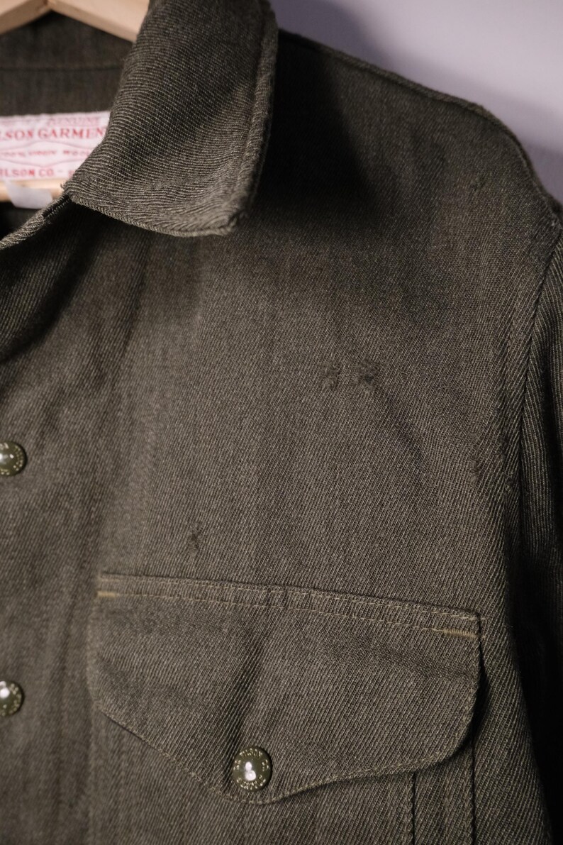 Puede incluir: Primer plano de una camisa verde oliva oscuro con dos bolsillos en el pecho y cierres de bot&oacute;n. La camisa tiene cuello y una etiqueta que dice "Wilson Garment Co."