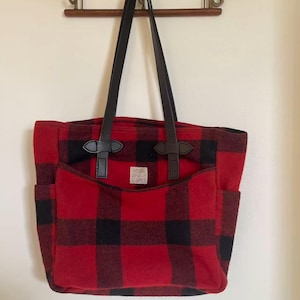 Op de afbeelding: Een rood-zwarte geruite tote bag met zwarte leren handvatten en accenten. De tas heeft een grote voorzak en twee zijzakken. Een klein label is zichtbaar op de voorzak.