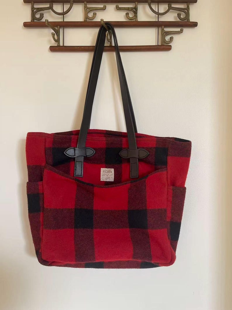 Op de afbeelding: Een rood-zwarte geruite tote bag met zwarte leren handvatten en accenten. De tas heeft een grote voorzak en twee zijzakken. Een klein label is zichtbaar op de voorzak.