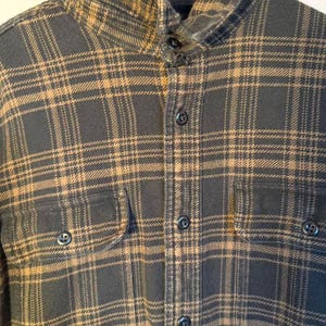 May include: A dark gray and tan plaid flannel shirt with a button-down front, two chest pockets, and a collared neckline. The shirt is hanging on a wooden hanger.