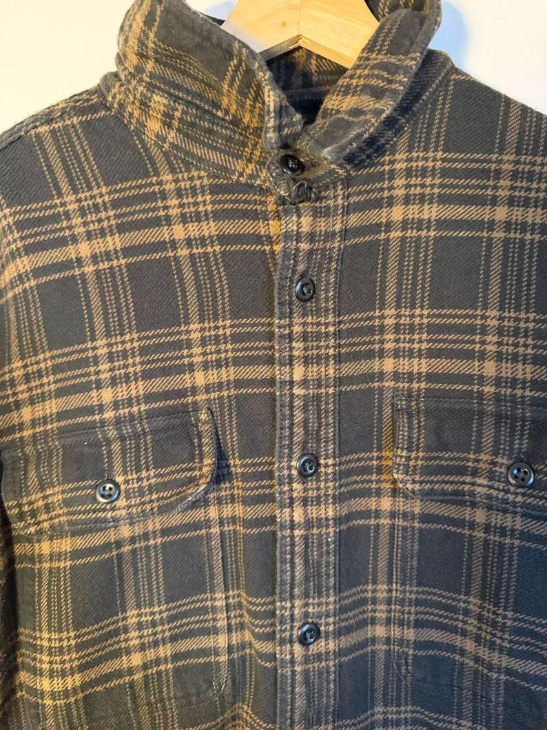 May include: A dark gray and tan plaid flannel shirt with a button-down front, two chest pockets, and a collared neckline. The shirt is hanging on a wooden hanger.