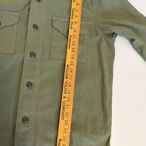 May include: Olive green button-up shirt with two chest pockets. The shirt has a collar and long sleeves. The brand name "FILSON" is visible on the bottom right. A yellow ruler is placed vertically on the shirt.
