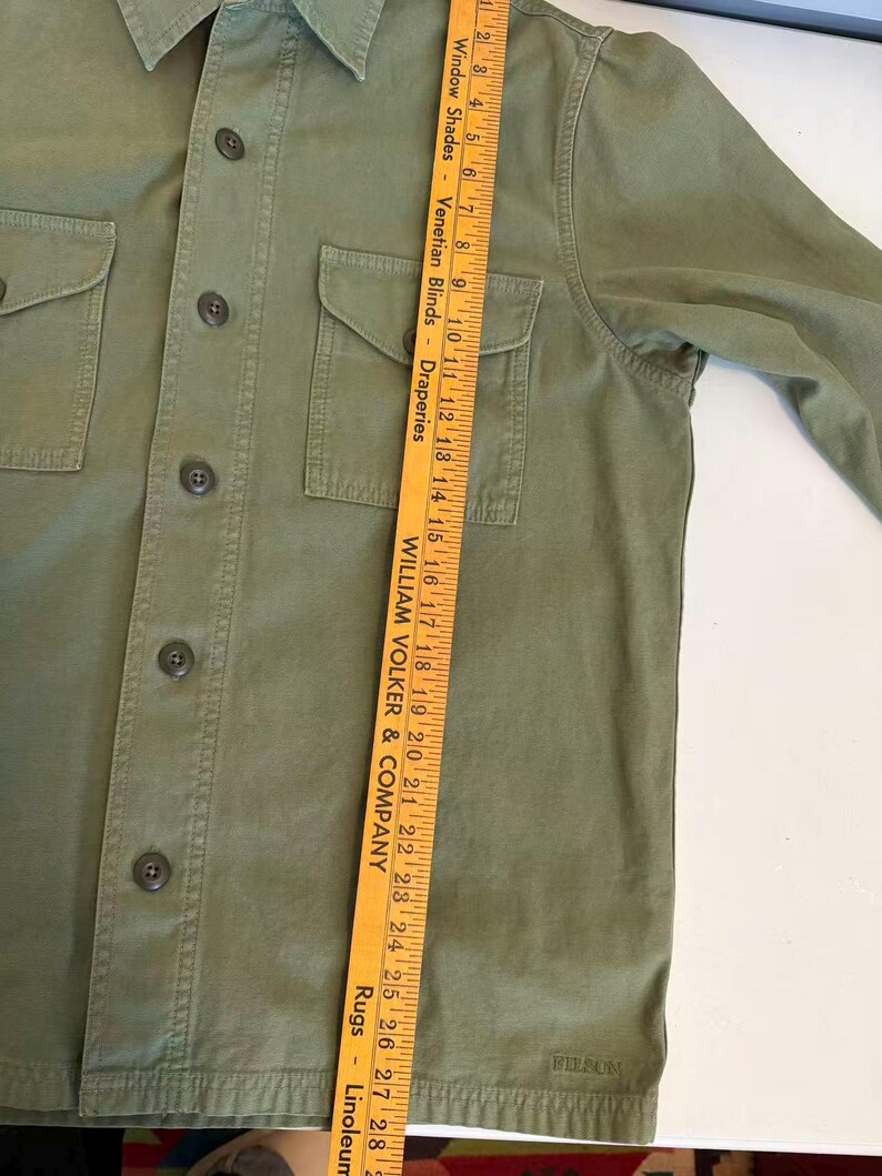 May include: Olive green button-up shirt with two chest pockets. The shirt has a collar and long sleeves. The brand name "FILSON" is visible on the bottom right. A yellow ruler is placed vertically on the shirt.