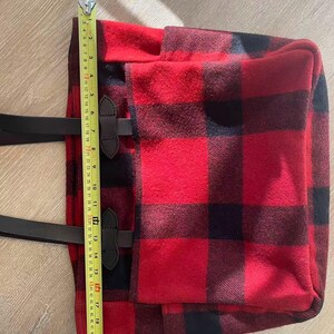 Op de afbeelding: Rood en zwart buffelruit draagtas met zwarte leren handvatten en accenten. De tas is ongeveer 48 cm hoog. De draagtas heeft een rechthoekige vorm en een klepsluiting met leren riemen en gespen. De tas is gemaakt van een dikke, geweven stof.