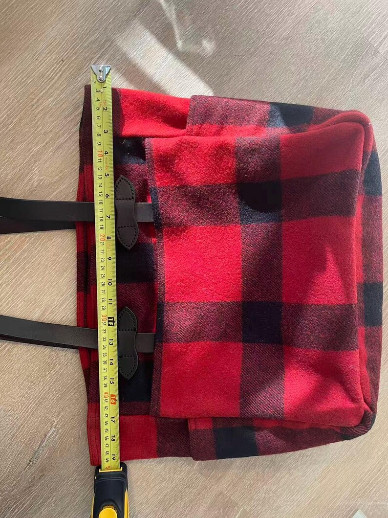 Op de afbeelding: Rood en zwart buffelruit draagtas met zwarte leren handvatten en accenten. De tas is ongeveer 48 cm hoog. De draagtas heeft een rechthoekige vorm en een klepsluiting met leren riemen en gespen. De tas is gemaakt van een dikke, geweven stof.