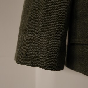 Puede incluir: Primer plano de una manga de chaqueta verde oscuro. La tela tiene un tejido diagonal texturizado. El pu&ntilde;o es visible, con unas peque&ntilde;as puntadas decorativas cerca del borde. La chaqueta parece estar hecha de un material duradero.